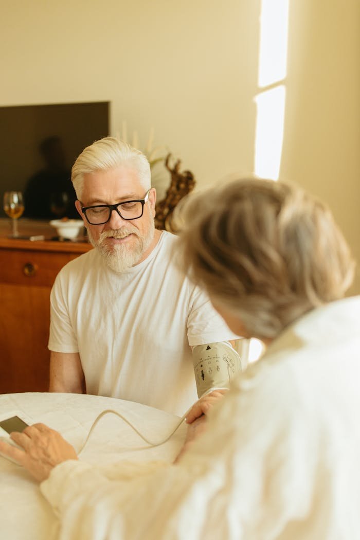 Senior man with gray hair and glasses checking blood pressure using a digital monitor at home.